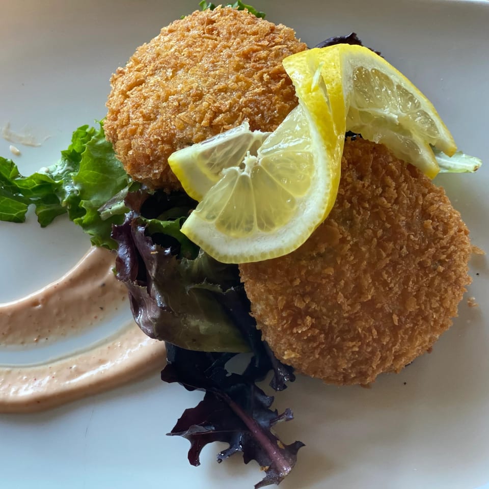 Two golden-brown crab cakes with lemon wedges, mixed greens, and a creamy sauce on a white plate.