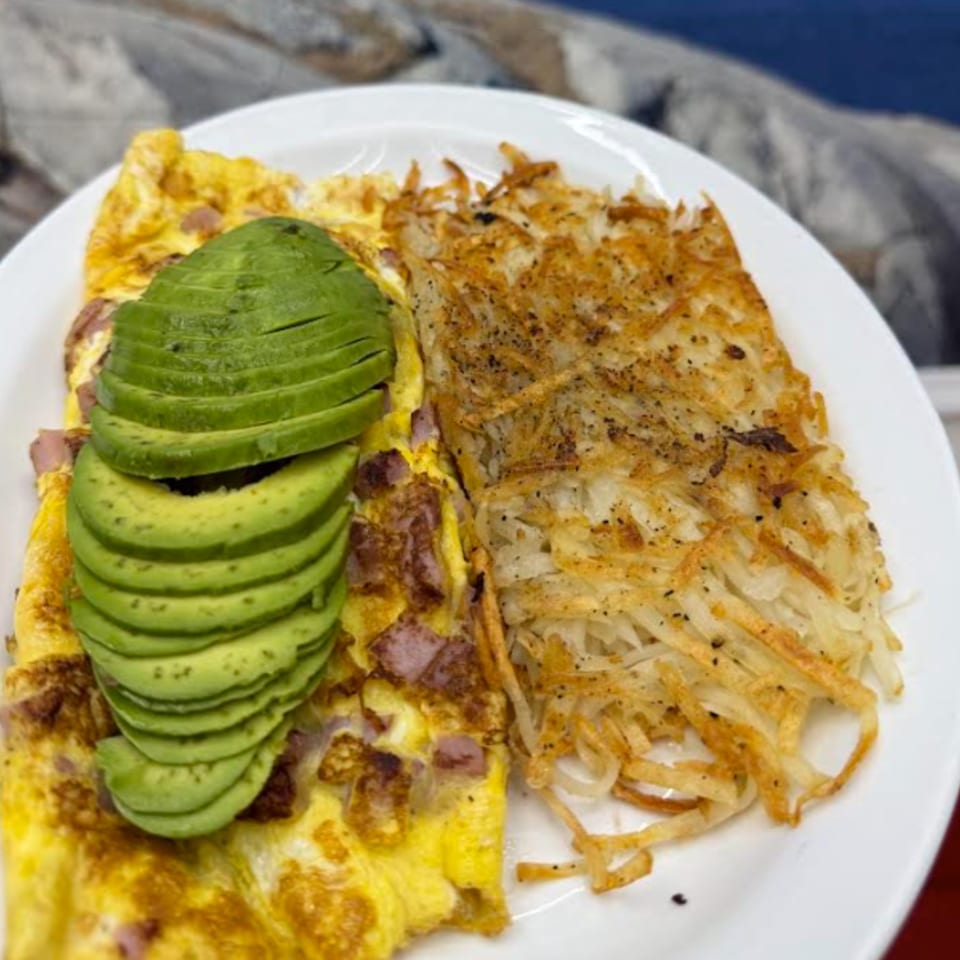 A breakfast plate with a ham and cheese omelet topped with sliced avocado, and crispy hash browns.